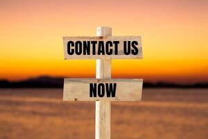 Contact us now message written on wooden sign with blurry lake background. Conceptual contact us now symbol. Copy space. photo
