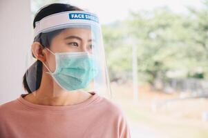 Asian woman wearing mask and face shield before going outside her room for prevent and protection covid-19 virus disease. Conceptual of new normal lifestyle after Covid-19 pandemic outbreak. photo