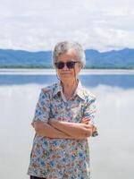Happy a senior woman with short gray hair smiling, arms crossed, and looking at the camera standing near lake and mountains background. Concept of aged people and relaxation photo