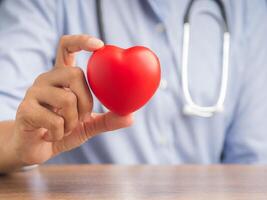 Hand of a doctor showing a red heart shape while sitting in the hospital or clinic. Close-up photo. Space for text. Medical and encouragement concept photo