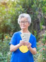 Cheerful senior woman with short gray hair holding US dollars banknote and looking at the camera with smile while standing in the garden photo