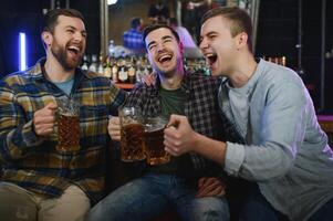 Three young men in casual clothes are smiling, holding bottles of beer while standing near bar counter in pub photo