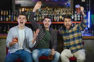 Soccer fans cheering for favourite team while enjoying drinks. Group of supporters celebrating victory while watching football match on tv in a club. Happy friends shouting while cheering for a goal. photo