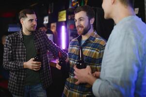 Three young men in casual clothes are smiling, holding bottles of beer while standing near bar counter in pub photo
