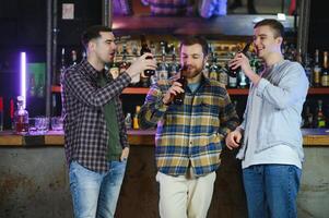 Three young men in casual clothes are smiling, holding bottles of beer while standing near bar counter in pub photo