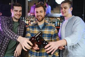 Three young men in casual clothes are smiling and clanging glasses of beer together while sitting at bar counter in pub photo