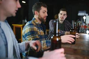 Three young men in casual clothes are smiling, holding bottles of beer while standing near bar counter in pub photo
