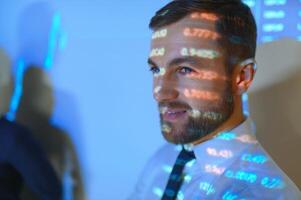 Stock Market Top Trader Looks at Projected Ticker Numbers and Graphs Running, Analysing Data to Make Best Sell. Behind Him Room Full of Screens and Statistics. photo