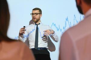 Businessman does Financial Analysis talks to Group of Businessspeople. Projector Screen Shows Stock Market Data, Investment Strategy, Revenue Growth photo