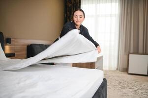 Young pretty housekeeper in uniform changing bedclothes in hotel room while standing by double bed photo