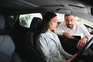 Satisfied happy caucasian female client customer woman sitting at the wheel of new car while male shop assistant helping her choose it photo