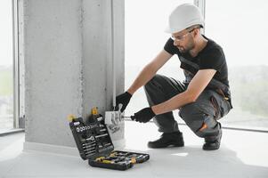 Confident professional electrician in uniform using screwdriver while replacing a socket in apartment after renovation work photo