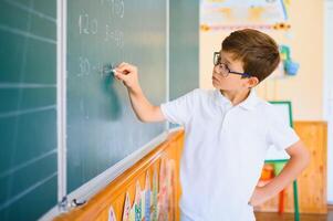 Cute little schoolboy diligent write chalk solve math problem dressed stylish uniform classroom blackboard background photo