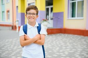 Portrait of little schoolboy in glasses and with backpacks standing on the school building. Study concept photo