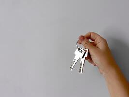 Close up of hand holding set of silver keys against plain light grey background with copy space. Minimalistic image symbolizing real estate, property ownership, or access with focus on metal keys. photo