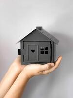 Close up of hands holding black house model with cut out heart and windows against grey background. Minimalistic, symbolic image representing real estate, home and property ownership. photo