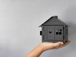 Close up of hand holding black house model with cut out heart and windows against grey background with copy space. Minimalistic, symbolic image representing real estate, home and property ownership. photo