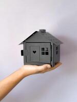 Close up of hand holding black house model with cut out heart and windows against grey background with copy space. Minimalistic, symbolic image representing real estate, home and property ownership. photo