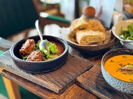 Close up of rustic wooden table with braised meat in tomato sauce garnished with cilantro, served in black bowl, along with crusty bread, creamy pumpkin soup, and mixed green salad in background. photo
