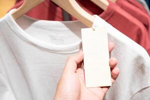 Hand holding blank paper price tag. blank tag with Clothes on a shelf in a designer clothes store. Blank for text. photo