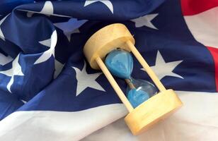 Hourglass on table with USA flag in the background. Sand glass on old wooden table with American flag in the background, vertical image. photo