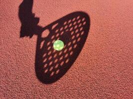 High angle of yellow ball placed on court and covered with shadow of crop person holding padel racket photo