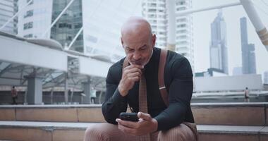 Manager sits in busy metropolis, smiling and staring at phone screen during work break. Man enthusiastically scrolls through social media feed on phone. Bustle city does not distract from phone. video