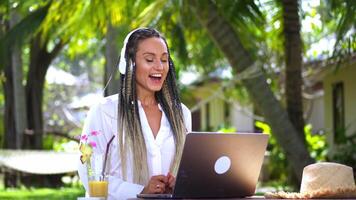 Woman tourist talking via web link call using laptop while sitting in tropical cafe while relaxing. Young woman tourist in headphones communicates via call using laptop at resort cafe. video