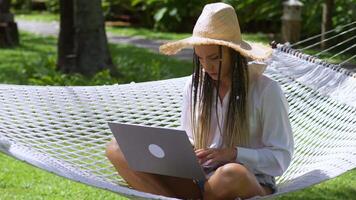 Female digital nomad in straw hat uses laptop to online working, while relaxing on hammock at summer vacation. Female digital nomad using laptop to freelance work while traveling. Remote work concept. video
