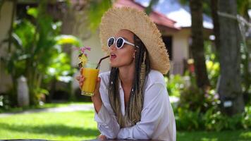 Portrait glad woman tourist enjoying summer cold drink banana fresh juice, resting on summer vacation. Woman in sunglasses and hat drinking orange juice at summertime. Concept resting on vacation video