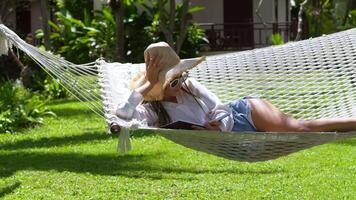Relaxed female tourist in straw hat lies on a hammock and reads a book on a hot summer day. Woman is resting on summer vacation in a tropical country, reading a book relaxing on a hammock at resort video