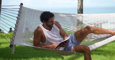 Attractive relaxed dark-haired multi-ethnic bearded man tourist reading a book, relaxing on a hammock, surrounded by a tropical beach with turquoise water and a green park. Handsome guy on vacations video