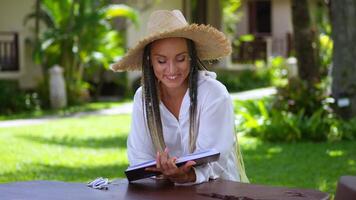 Young woman in straw hat enthusiastically reads book and smiles during summer vacation in tropics on background of palm trees at local outdoors cafe table. Breeze from sea shakes the straw hat video