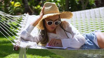 Woman tourist in straw hat resting hammock and reading book at summer holidays. Female tourist resting on summer holidays in tropical country, reading book relaxing on hammock at tropical resort video