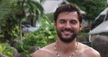 Headshot of an attractive charming young Caucasian bearded macho man, standing with naked torso against tropical nature background, enjoying sunbathing, smiling a beautiful toothy smile, at the camera video
