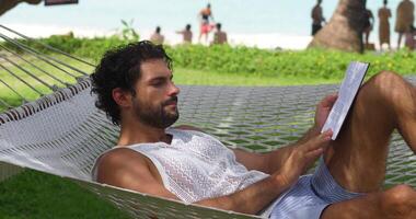 atractivo joven barbado masculino turista, disfruta verano Días festivos a el playa, siesta, acostado en un hamaca y leyendo un libro en un calentar soleado día. conocimiento. erudición. digital desintoxicación video