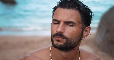 Close-up portrait. Attractive bearded Caucasian man, with wet body, after swimming on the tropical beach, enjoying sunbathing on a sunny day, standing against ocean background, looking at the camera video