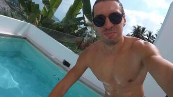Funny man enjoying vacation in tropical country filming himself on camera as he dives into the pool while enjoying summer holidays. Male in glasses jumping at pool on vacations. Selfie POV shot video