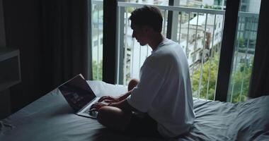Remote software developer, working diligently on his laptop while comfortably seated on home bed, crafting intricate lines of programming code. Back view male programmer telecommuting from his bedroom video