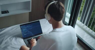 Rear view man freelancer programmer in headphones sitting on bed in bedroom and working remotely on laptop. Freelance man type code and programming on screen laptop, working from home video