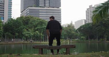 Young 30s man sits on a park bench against the backdrop of a lake in a city park outdoors. Lonely man walks in the park, rests on a bench in the park on backdrop of a lake and buildings in big city video