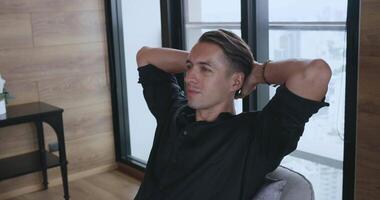 Close up serene calm millennial man in black shirt put hands behind head lean on comfy chair resting alone at home in front of window with big city view. 30s man enjoy fresh conditioned air indoors video