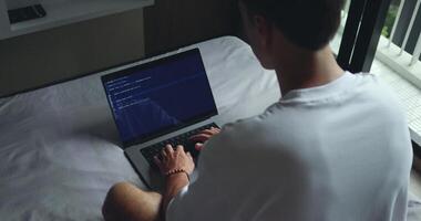 digital área de trabalho. homem digitando programador código em computador portátil, sentado em cama, traseiro visualizar. isto trabalhador autonomo escrevendo código em dele computador portátil a partir de a conforto do dele cama video