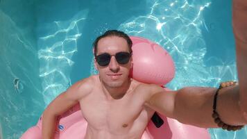 Happy man in sunglasses enjoying summer vacation filming himself swimming on a pink flamingo in a swimming pool during summer time video