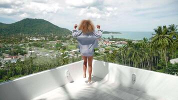 View from the back to a young redhead curly-haired European woman, running along the summer terrace of his apartment, overlooking the seaside and raising up hands, enjoying freedom and happiness video