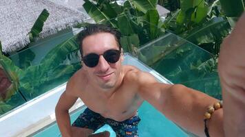 Young man glasses jumps into the pool during summer holidays in a tropical country. Millennial man enjoys an active summer holiday in the pool of hotel at summer vacations video