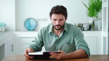 Handsome young Caucasian man sitting at a wooden table in the home kitchen interior, enjoying reading book. Charming guy relaxing while reading novel at his weekend. Leisure Hobby Literature Erudition video