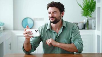 Attractive dark-haired bearded male freelancer, having a conference, communicating with his coworkers by call on smartphone, smiling looking at web camera while working remotely from home video