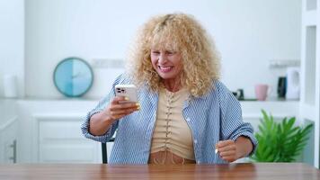 Angry blonde curly-haired caucasian woman using smartphone, typing text, scrolling news feed, experiencing rage and anger, feeling anxious, enraged, reading worst news and answers to a shocked message video