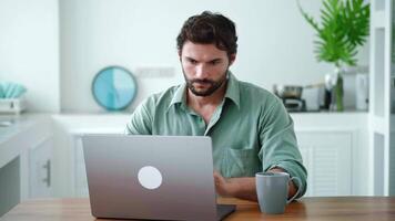 Front view of a handsome young freelancer working with laptop computer remotely, sitting at wooden table in kitchen. Pleasant happy man communicating in social network, searching information online. video